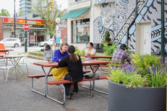 Two people sit at a picnic table on a side street in Vancouver's South Granville neighbourhood. There is a large mural on the building behind them. 