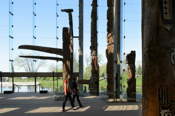 Vistors walk past totem poles and a large glass wall at the Museum of Anthropology in Vancouver