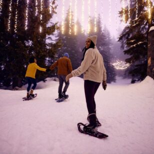 Three people wear snowshoes on a snowy path lit with lights in the forest at Grouse Mountain