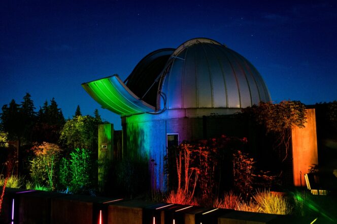 The exterior of the SFU Trottier Observatory in Vancouver at night with the telescope door open and the building lit up with soft colourful lights