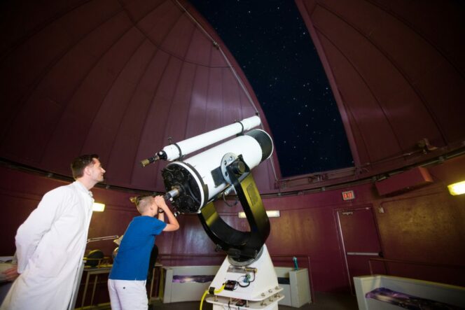 A scientist stands next to a boy who is looking through a giant telescope in the GMS Observatory at the HR MacMillan Space Centre in Vancouver